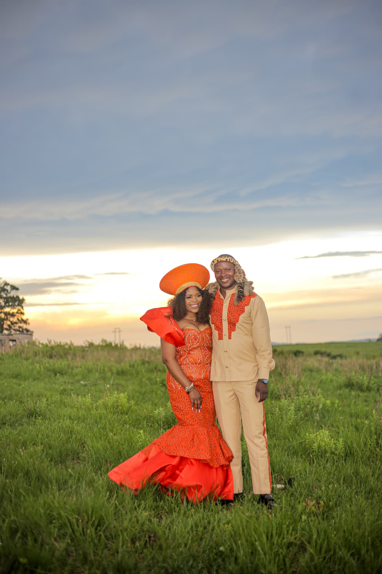 Couple in orange Zulu traditional wedding attire standing in a grass field at sunset