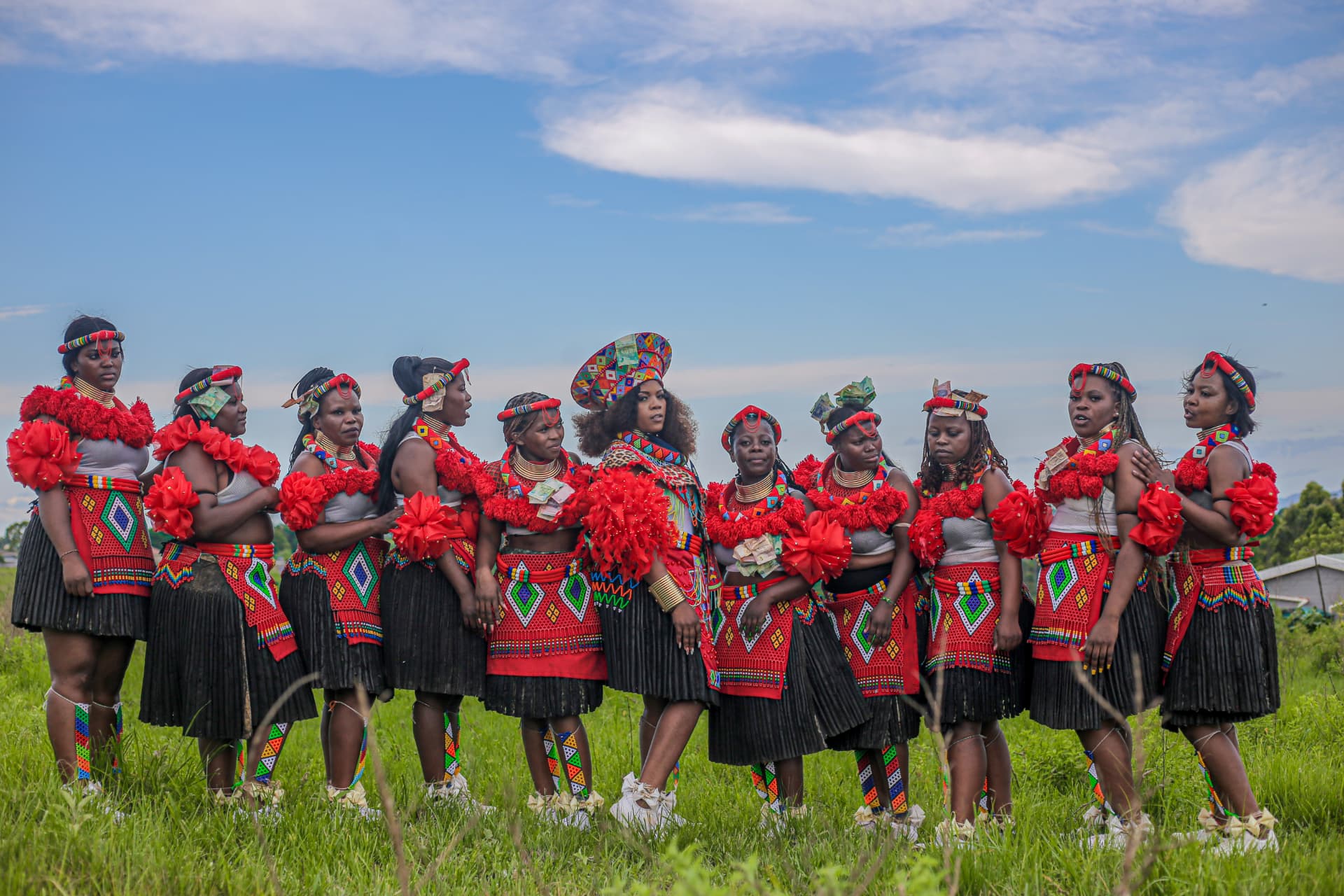 Row of women in red and black Zulu regalia standing in a green field under open sky